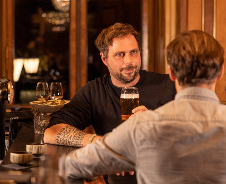 Zwei Männer unterhalten sich an der Bar, einer hält ein Glas Bier, die Atmosphäre ist warm und entspannt.